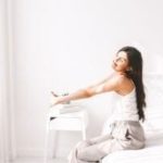 Young woman in casual clothing stretching in a minimalistic bright bedroom, promoting home comfort and peace.