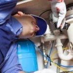 Plumber inspecting plumbing fixtures under sink, wearing gloves and cap, from B & D Plumbing, Heating & A/C.