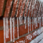 Icicles hanging from a textured brick wall illustrating winter challenges for plumbing, highlighting frozen pipe prevention.