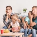 Family watching a scary movie on a couch, showing surprise; bowl of popcorn and drink on the table. B & D HVAC services.
