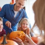 Man and two children carving pumpkins with a knife in a festive autumn home setting, promoting family activities.
