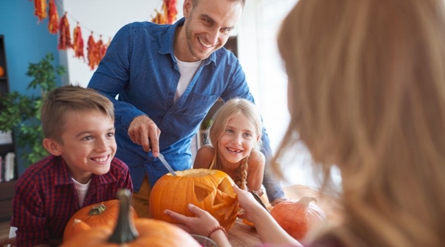 Smiling man and children carving pumpkins at a table, showcasing autumn family bonding and celebration.