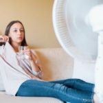 Girl sitting on a couch with a glass of water and an electric fan, highlighting comfort in HVAC service support.