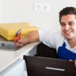 Smiling male technician in blue apron holding a wrench in a clean kitchen, promoting plumbing services by B & D Plumbing.