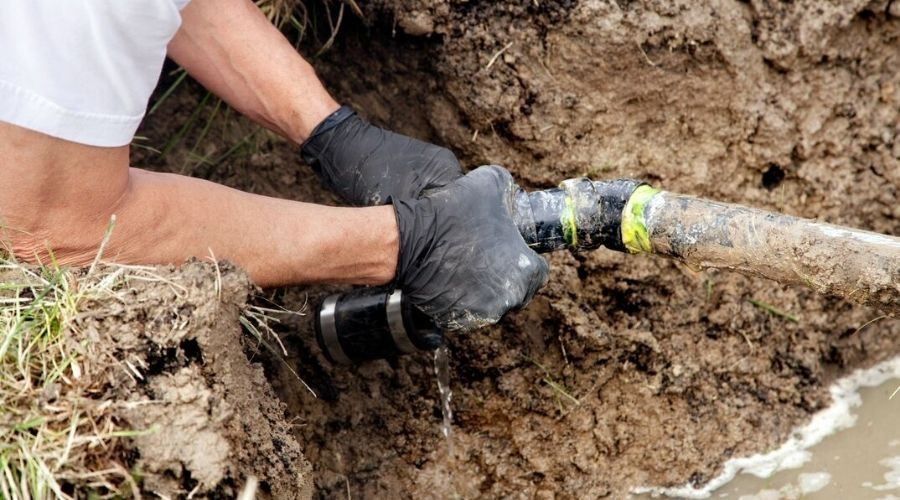 Person repairing a leaking pipe, wearing gloves, with excavated dirt and visible water, by B & D Plumbing.