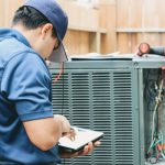 HVAC technician in uniform servicing an air conditioning unit with a tablet for B & D Plumbing, Heating & A/C.