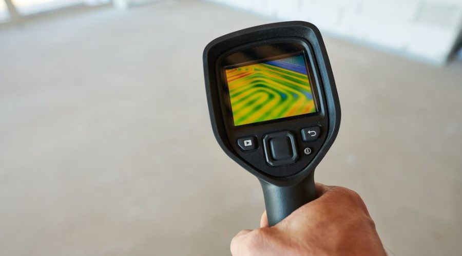 Thermal imaging camera held by a technician, showing colorful heat patterns in an indoor space with concrete flooring.