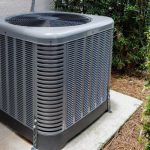 Air conditioning unit on concrete slab with gray casing, surrounded by greenery, by B & D Plumbing, Heating & A/C.