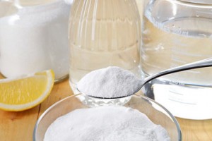 Baking soda in a bowl with a spoon, alongside vinegar, water, and a lemon slice, illustrating effective cleaning agents for deodorizing garbage disposals.